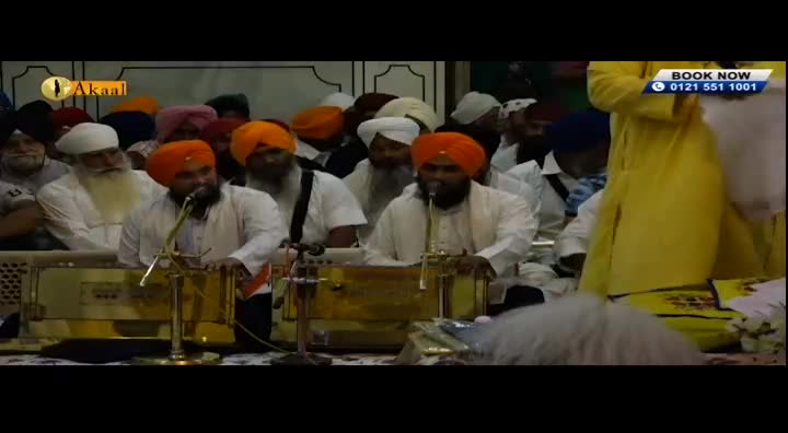 Two men in orange turbans are playing harmoniums, facing microphones. A crowd of people, many also wearing turbans, watches them from behind.
