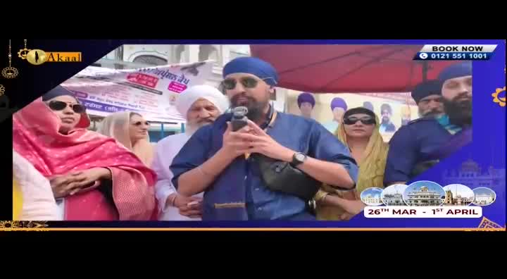 A man in a blue turban and shirt holds a microphone, speaking to a crowd gathered outdoors. Behind him, people in traditional attire watch, some with their heads covered.