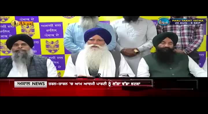 Three men sit behind a table, a vibrant yellow and purple banner with Sikh symbols behind them. A news ticker scrolls across the bottom of the screen, displaying text in Punjabi.
