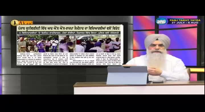 A man in a white turban and beard sits at a desk, looking towards the camera. On a screen behind him, a newspaper article with photos of a crowd and police is displayed, with the Akaal Channel logo visible.