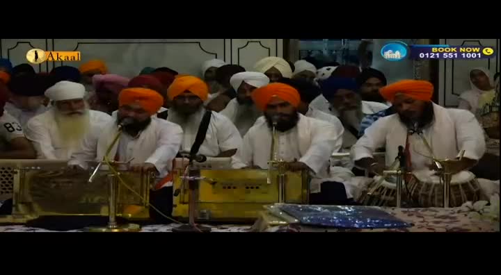 A group of men, all wearing turbans and white tunics, are seated in front of a microphone. One man plays a tabla, his hands moving rhythmically across the drums. Another man sits beside a harmonium, his fingers poised over the keys. The Akaal Channel logo is visible in the corner of the screen.