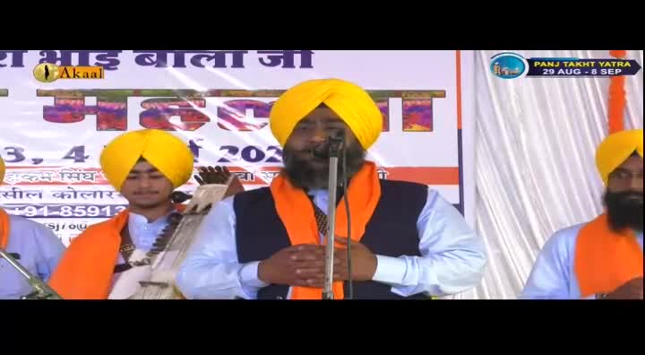 A man in a yellow turban sings into a microphone, his hands clasped. To his left, a young man plays a stringed instrument.