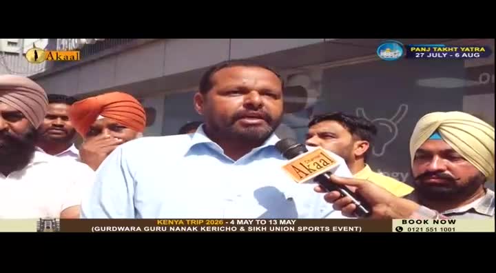 A man with a beard and a light blue shirt speaks into an Akaal Channel microphone. He stands surrounded by other men, some wearing turbans, in front of a building.