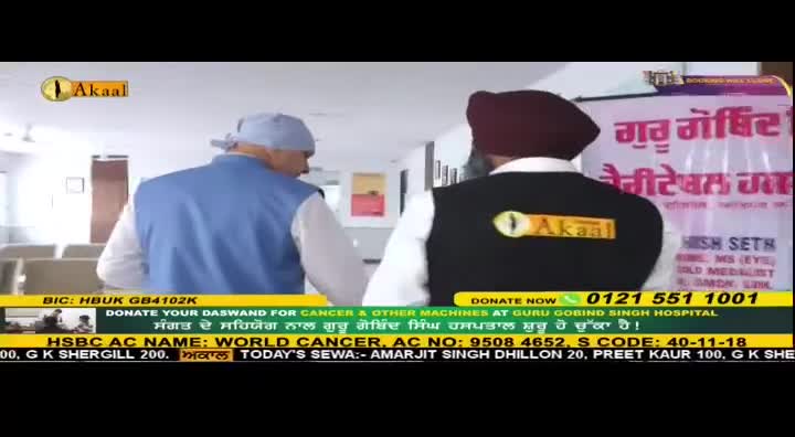 Two men walk down a hallway, one in a blue vest and cap, the other in a black vest with the Akaal logo. A banner behind them announces "Guru Gobind Singh Hospital."