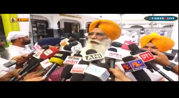 A man in an orange turban speaks into a cluster of microphones, his long white beard prominent. Several other men, also wearing turbans, stand close by, holding microphones and cameras.