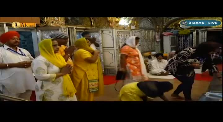 A group of people, some in bright yellow, stand with hands clasped in prayer. A woman in a black and white patterned top bends low, her head bowed. This is being broadcast live on Akaal Channel from the UK.