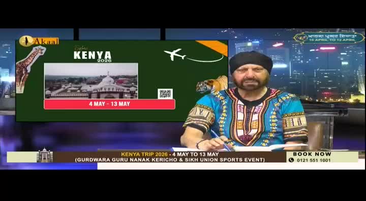 A man in a vibrant blue and gold patterned shirt sits at a desk, looking directly ahead. Behind him, a screen displays a giraffe, an airplane, and text promoting a "Kenya Trip 2026."