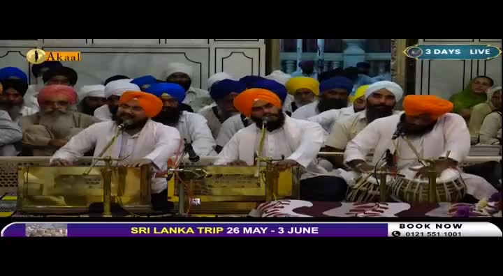 A group of men, all wearing turbans, are gathered. Two men in the foreground are playing musical instruments, one on a harmonium and the other on a tabla. The Akaal Channel logo is visible in the upper left corner.