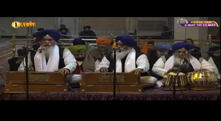 Four men, dressed in traditional Sikh attire, are seated on a raised platform, playing musical instruments. Two men are playing harmoniums, while another is playing a tabla, all singing into microphones. The Akaal Channel logo is visible in the top left corner.