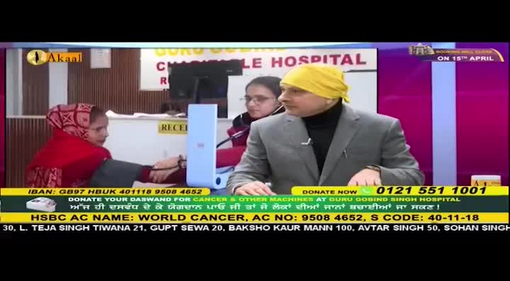 A man in a grey suit and yellow headscarf speaks into a microphone, while a woman in a red head covering sits opposite him at a reception desk. A screen behind them displays "GURU GOBIND SINGH HOSPITAL" and the Akaal Channel logo.