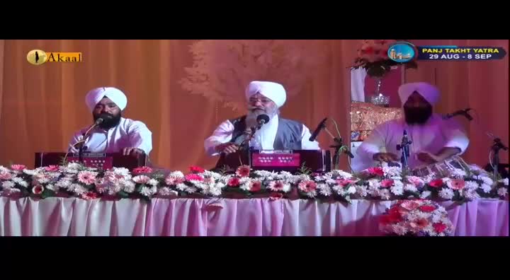 Three men in white turbans are seated behind a table adorned with flowers. The man in the center sings into a microphone while playing a harmonium, and the man on the right plays a tabla. This performance is being broadcast by Akaal Channel.