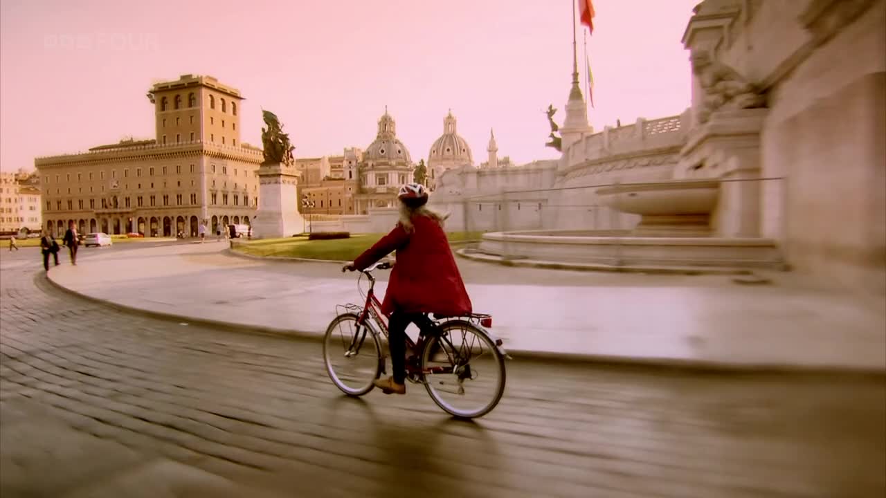 A cyclist in a red coat pedals across the cobblestone. The camera follows them, showing a backdrop of grand buildings and a pink sky.

