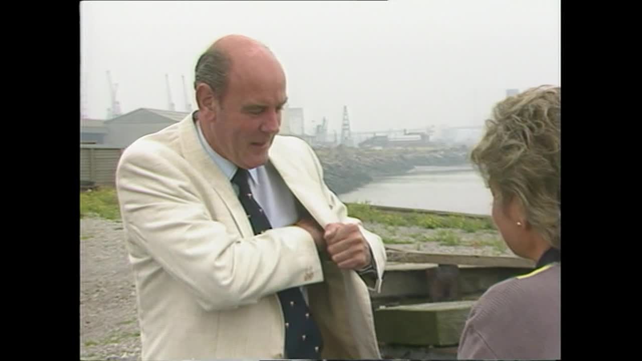A man in a cream blazer reaches into his jacket pocket, looking down as he does so. Across from him, a woman watches, the industrial backdrop of the United Kingdom visible behind them.
