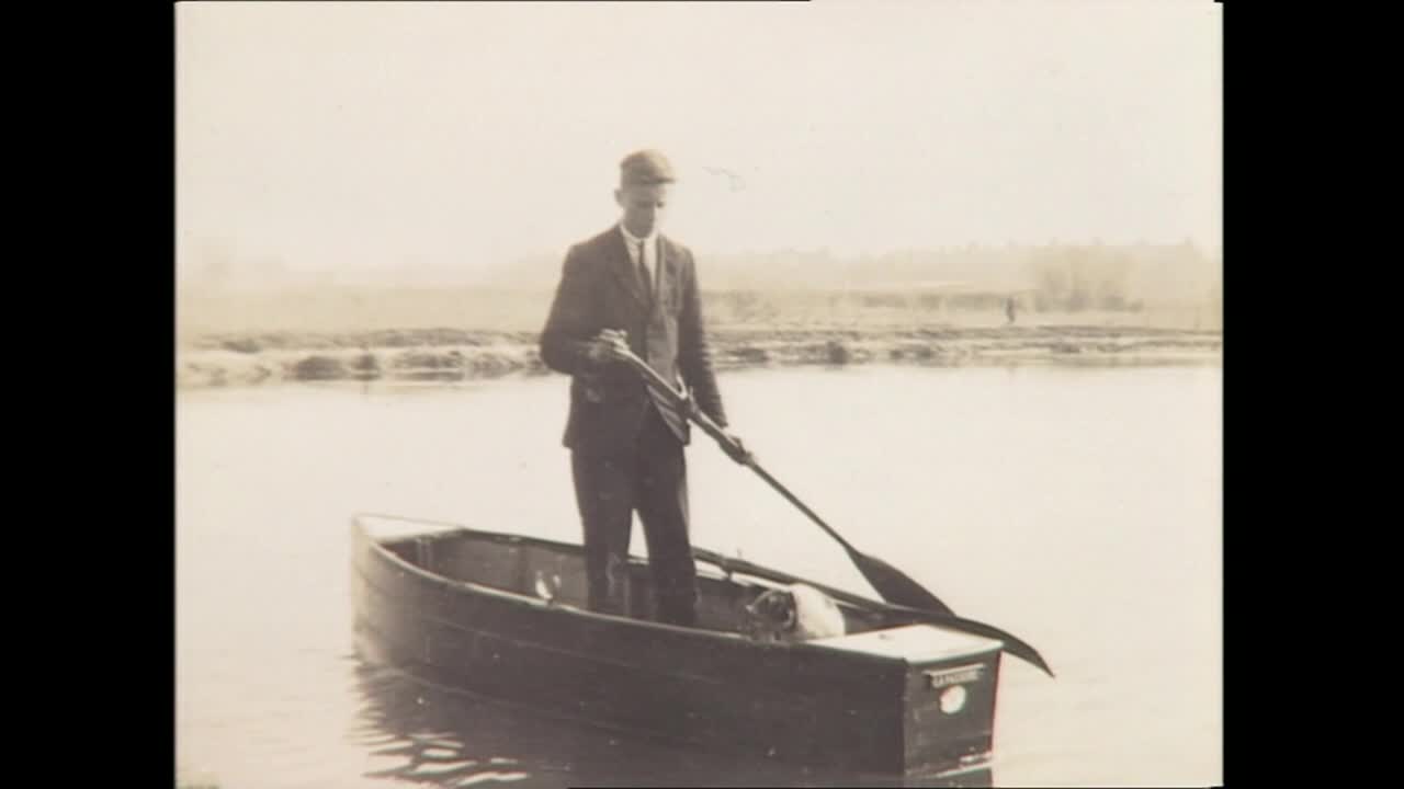 A man in a suit stands in a small boat, holding a paddle and a long gun. He's in the middle of the water, with a dog and a box in the boat with him.
