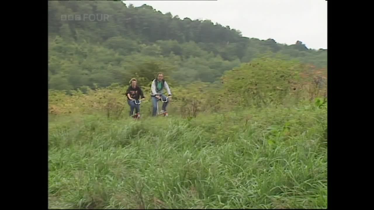 Two people are gliding along on scooters through tall grass, heading up a slight incline. The BBC Four logo is visible in the corner, and the background is a hillside covered in green.
