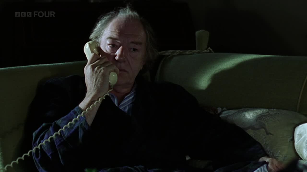 A man, wearing a dark jacket over a striped shirt, holds a cream-colored phone to his ear.  He's sitting on a green sofa, the BBC Four logo visible in the corner.
