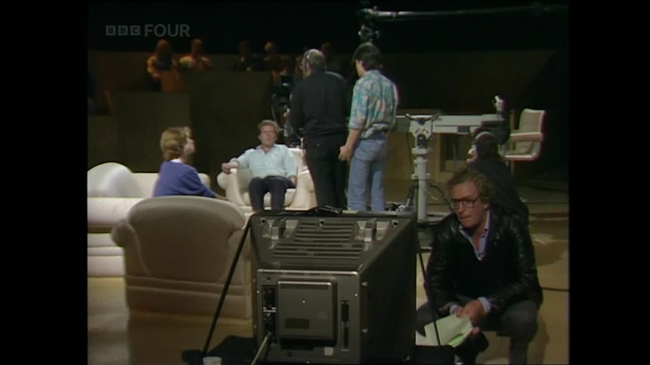 On a BBC Four set, two people sit in armchairs while crew members mill about. A man in a leather jacket kneels, looking at papers, as a camera monitor looms in the foreground.
