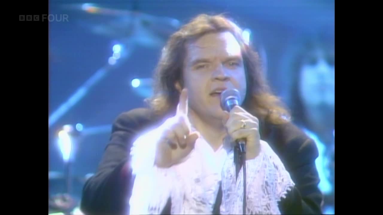 The singer, clad in a white, frilly shirt, points emphatically while holding a microphone. The BBC Four logo is visible, and the stage is awash in blue light.
