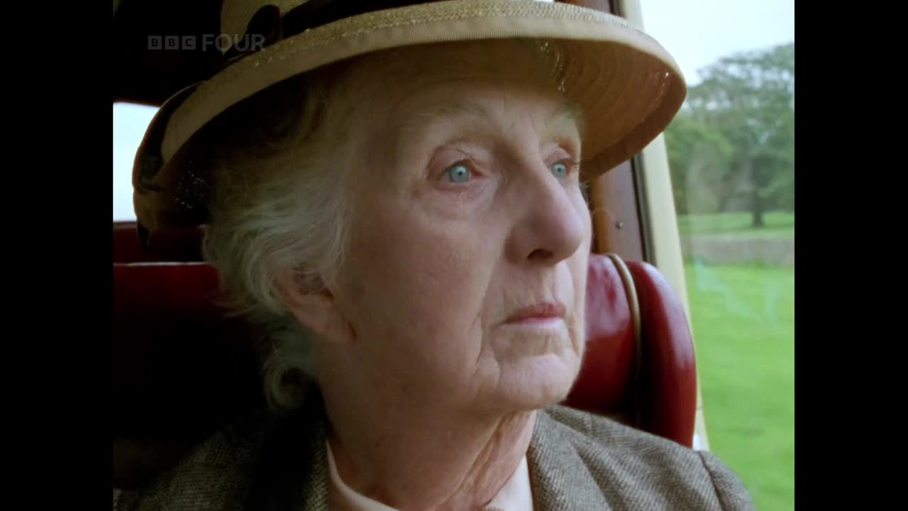 An older woman, wearing a hat, gazes out the window of a vehicle, her expression thoughtful. Outside, a green landscape rolls by, visible through the glass.
