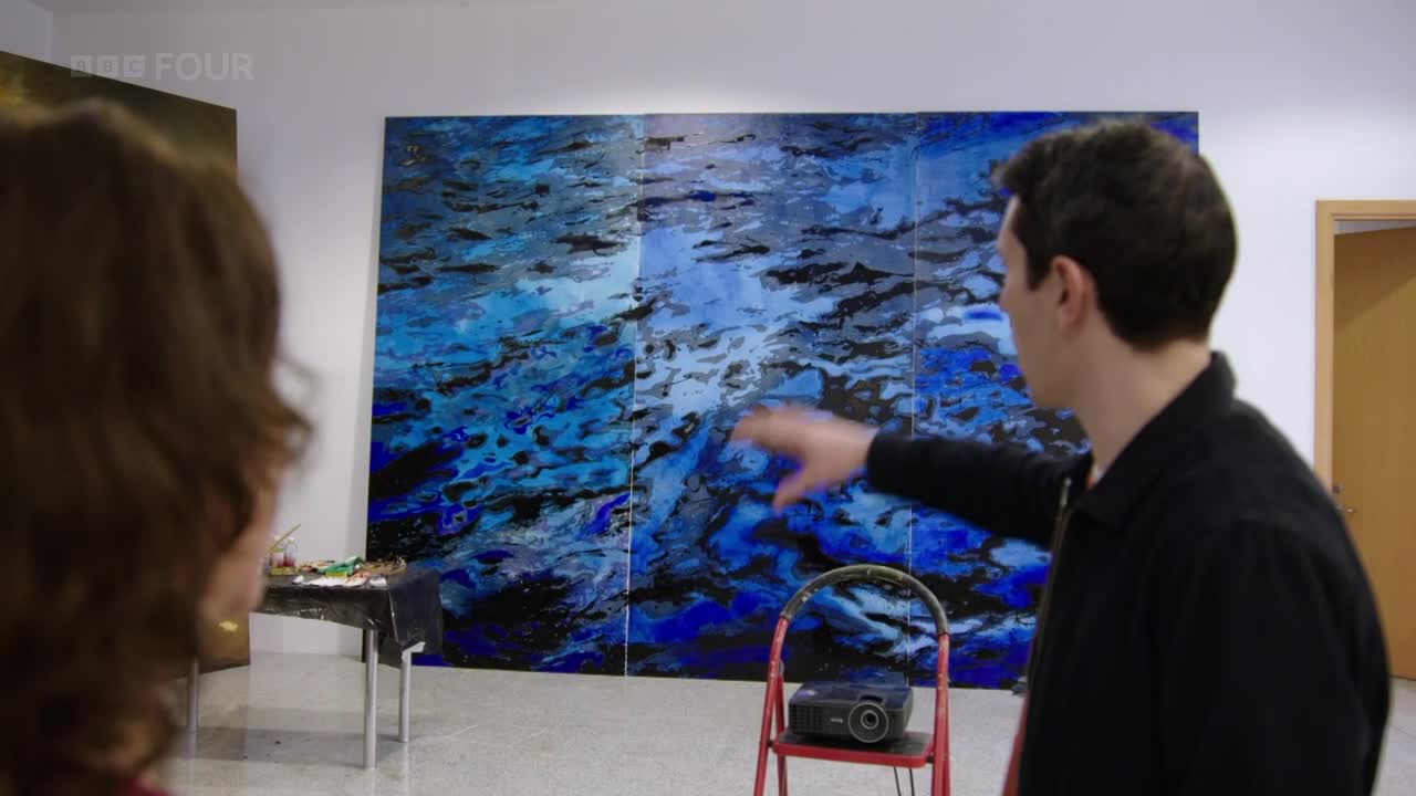 A man gestures towards a large, abstract painting of blues and blacks, likely explaining it to the person in front of him. A projector sits on a red step ladder, presumably set up for a presentation.
