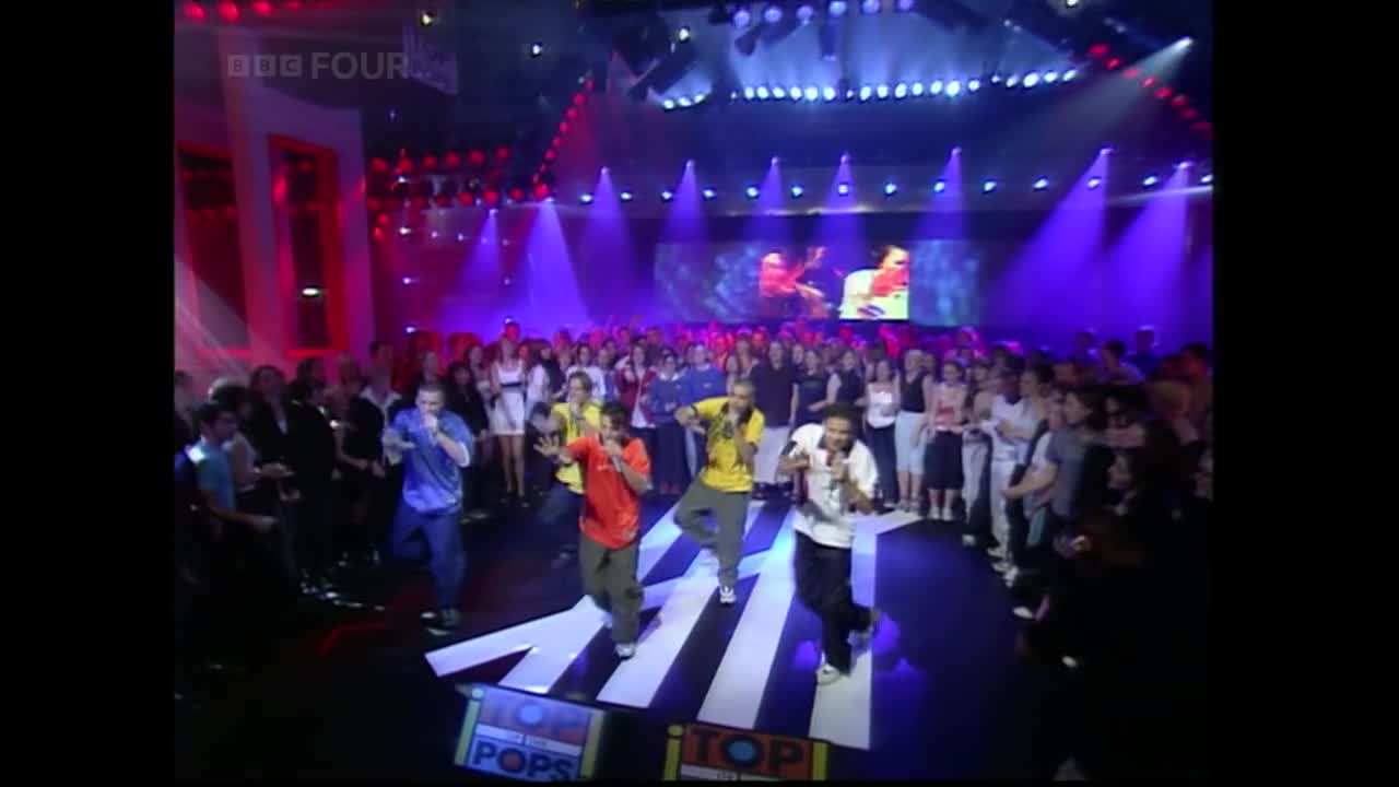On the BBC Four stage, a group of singers in bright shirts are performing, moving in sync on a giant, white-striped arrow. The crowd is clapping along, lit by purple and red stage lights.
