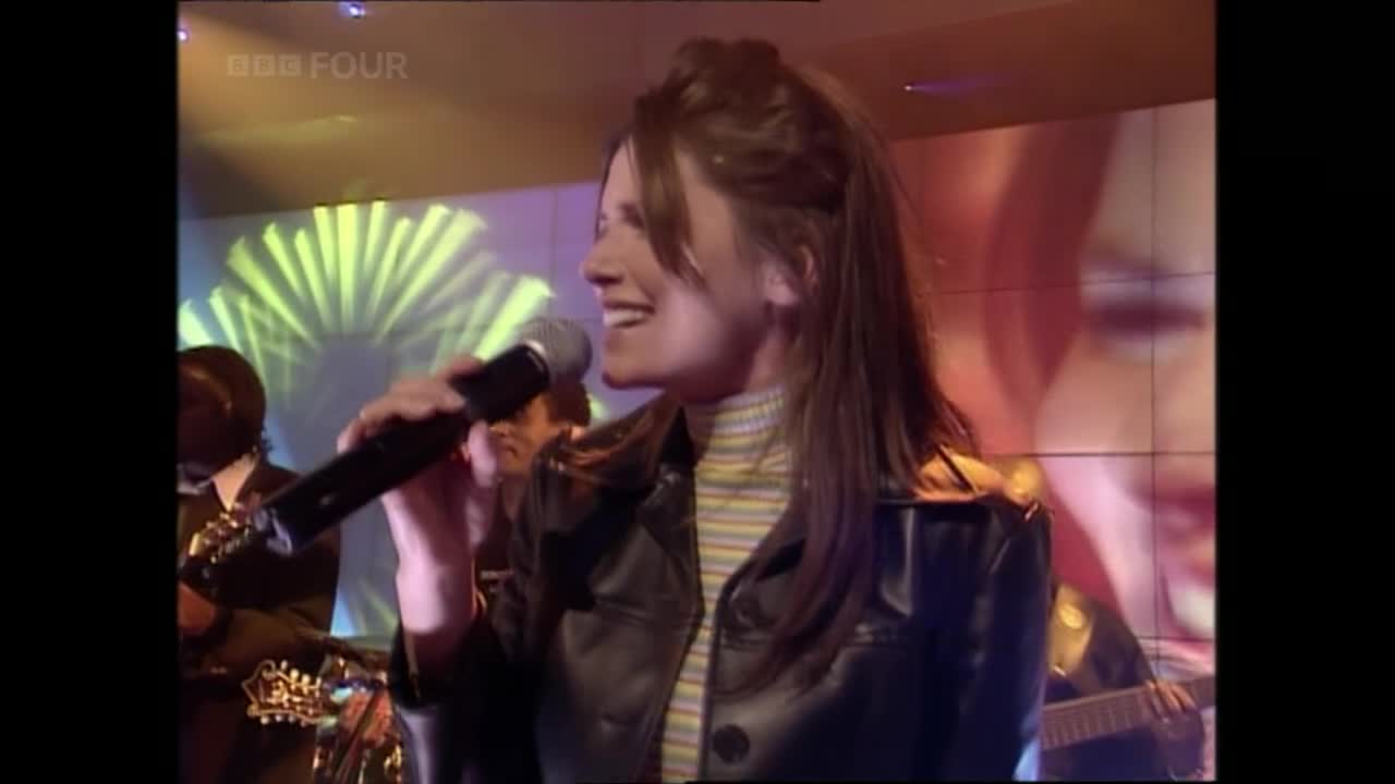 A woman in a leather jacket sings into a microphone, her face lit up. Behind her, a band plays on a BBC Four stage, the backdrop a swirl of light.
