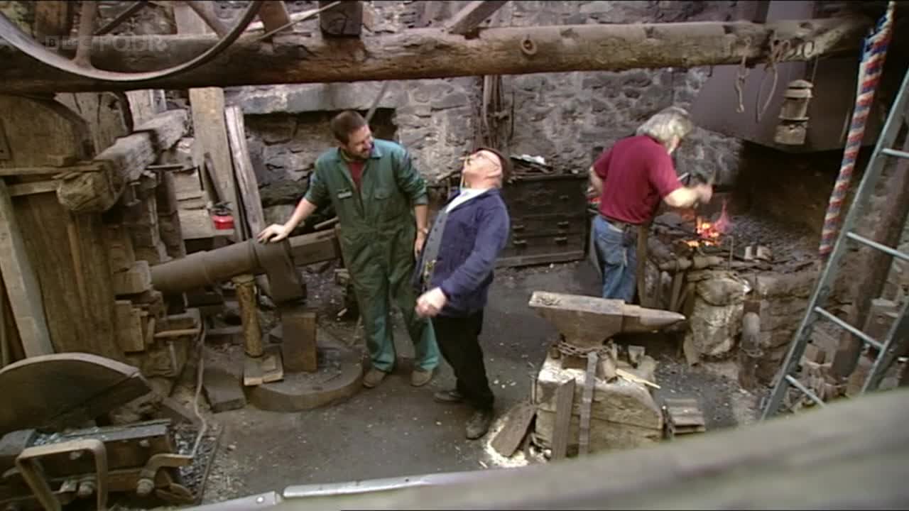 A man in a red shirt is working the forge, hammering something glowing red. Two other men watch, one in green overalls, the other with his head thrown back.
