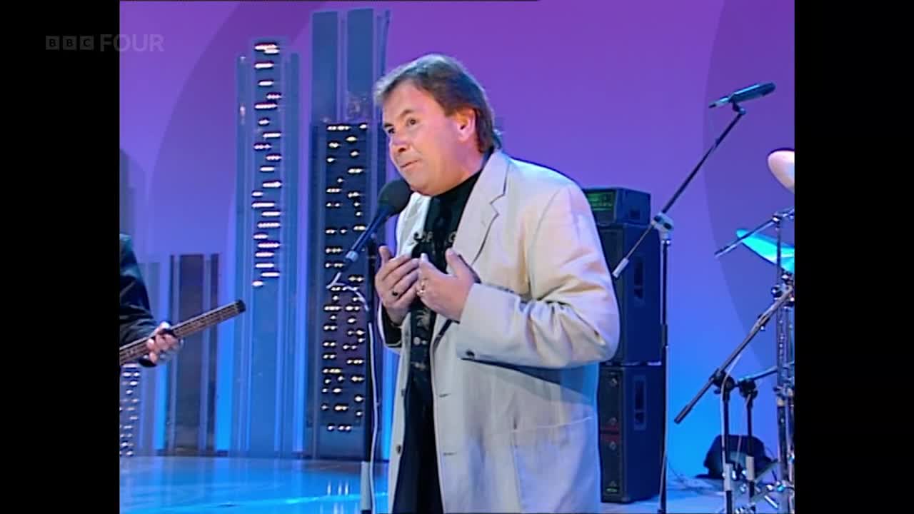 The singer, dressed in a light jacket, holds the microphone and gestures with his hands. Behind him, a band is set up on a stage, ready to perform on BBC Four in the United Kingdom.
The singer, dressed in a light jacket, holds the microphone and gestures with his hands. Behind him, a band is set up on a stage, ready to perform on BBC Four in the United Kingdom.