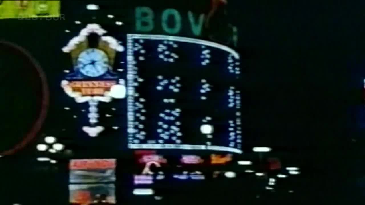 A giant clock face, lit up with neon lights, tells the time above a sign that reads "GREENWICH TIME". The word "BOV" is visible in green, and a grid of lights flickers on a large, curved screen.
