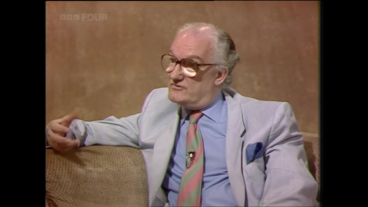 He's sitting on a sofa, gesturing with his left hand as he speaks. His tie has pink and green stripes, and the BBC Four logo is visible in the corner.
