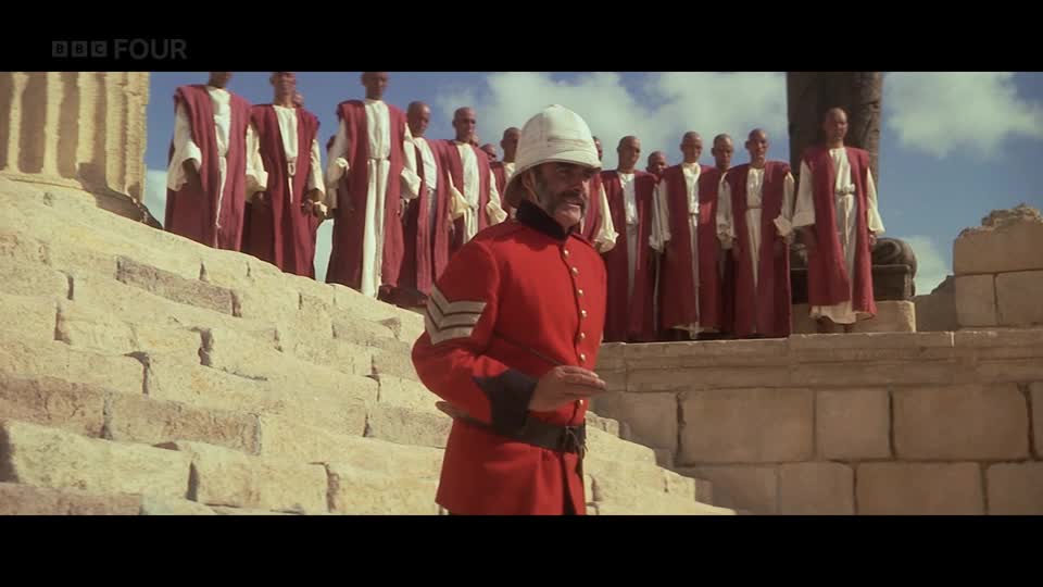A man in a red military uniform with a pith helmet stands on stone steps, facing a group of figures in red robes. The BBC Four logo is visible in the top left corner.