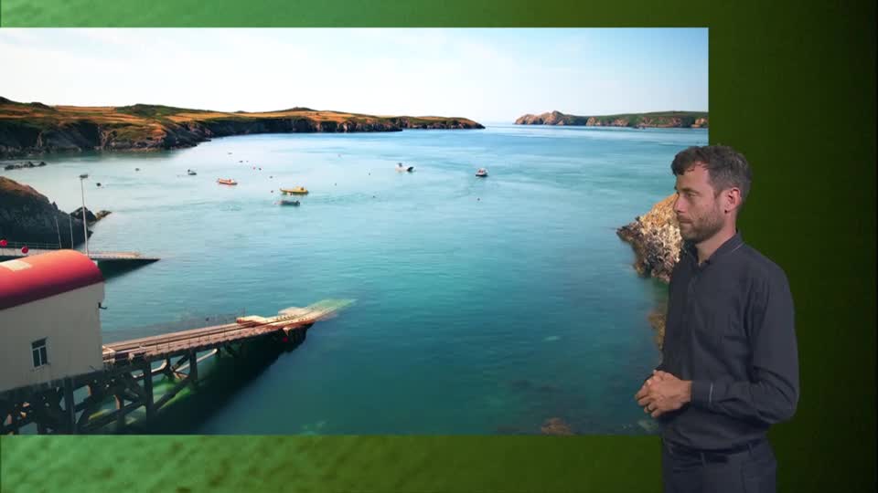 A man in a dark shirt stands beside a large screen displaying a picturesque Welsh bay. Small boats dot the clear blue water, leading towards rugged, sun-drenched cliffs.