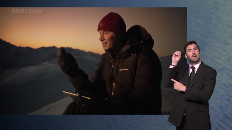 A man in a red beanie and black jacket holds a tablet, looking out at a snowy landscape as the sun sets. Beside him, a man in a suit signs with his hands.