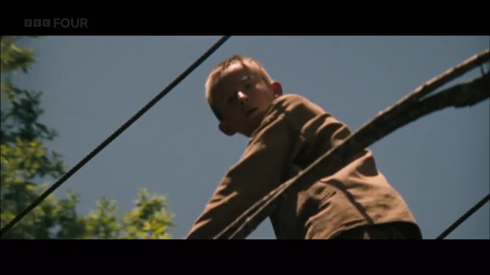 A young boy in a brown shirt looks up, his face a mask of apprehension. He's perched precariously on what appears to be a wooden structure, perhaps a treehouse, with thick ropes stretching across the frame.
