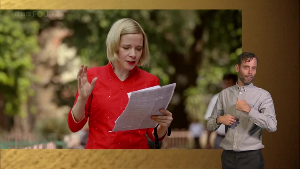 A woman in a red dress reads from papers, her hands gesturing. Beside her, a man in a grey shirt signs with his hands. This is part of a BBC Four broadcast from the UK.