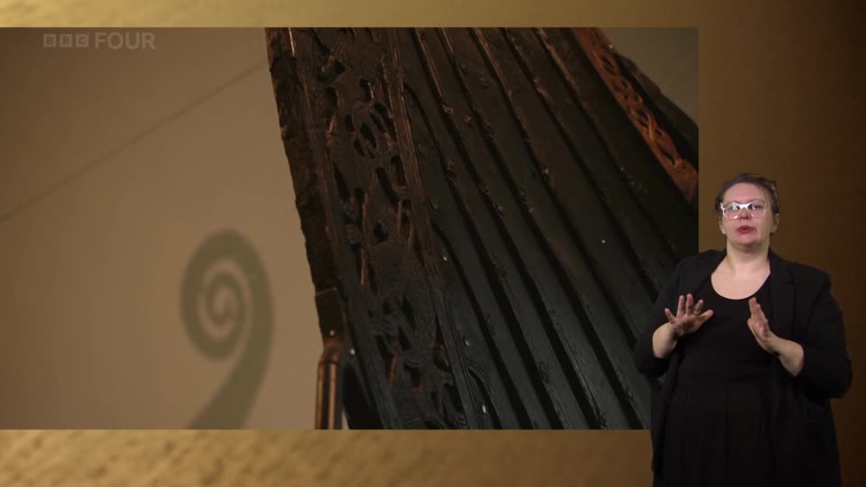 A woman is signing on BBC Four, her hands gesturing as a dark, carved wooden structure, possibly part of a ship, is displayed. A spiral shadow is cast on the wall behind the artifact.