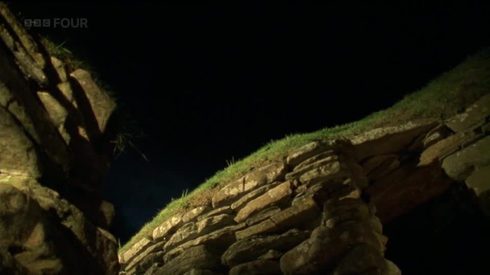 The drystone walls of an ancient structure are illuminated against the dark sky. A patch of bright green grass tops the rough-hewn stones, hinting at the rugged landscape of the UK.