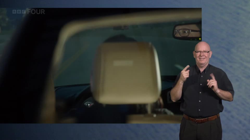 A man in a black shirt signs with his hands, his glasses reflecting the car interior. The BBC Four logo appears in the top left corner of the screen.
