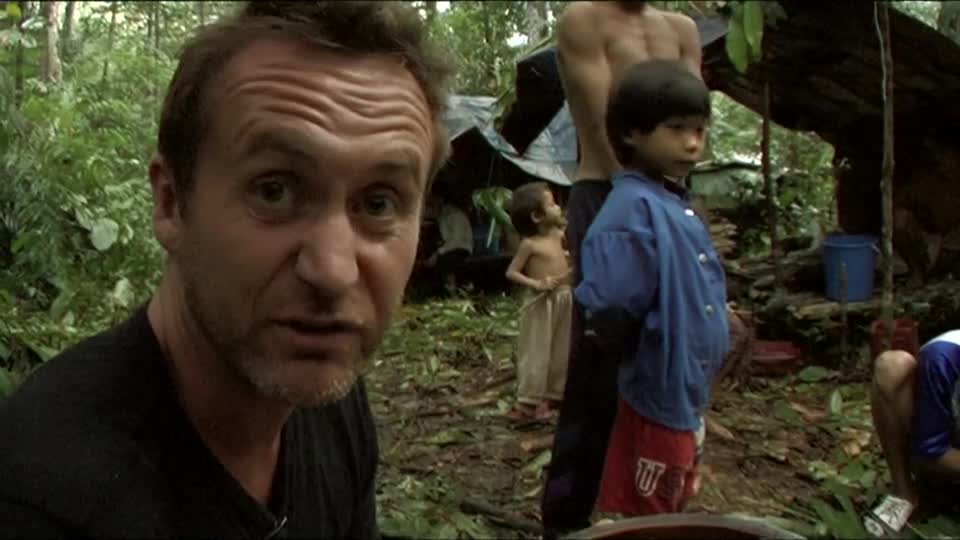 The man in the foreground is speaking directly to the camera, his face etched with expression. Behind him, a young boy in a blue shirt and red shorts stands attentively, surrounded by the dense green of the jungle.