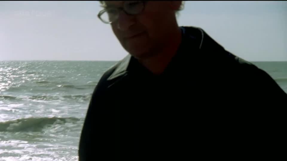 The sea is choppy under a pale sky, its surface glinting. A man in a dark jacket stands close to the camera, his glasses reflecting the light.