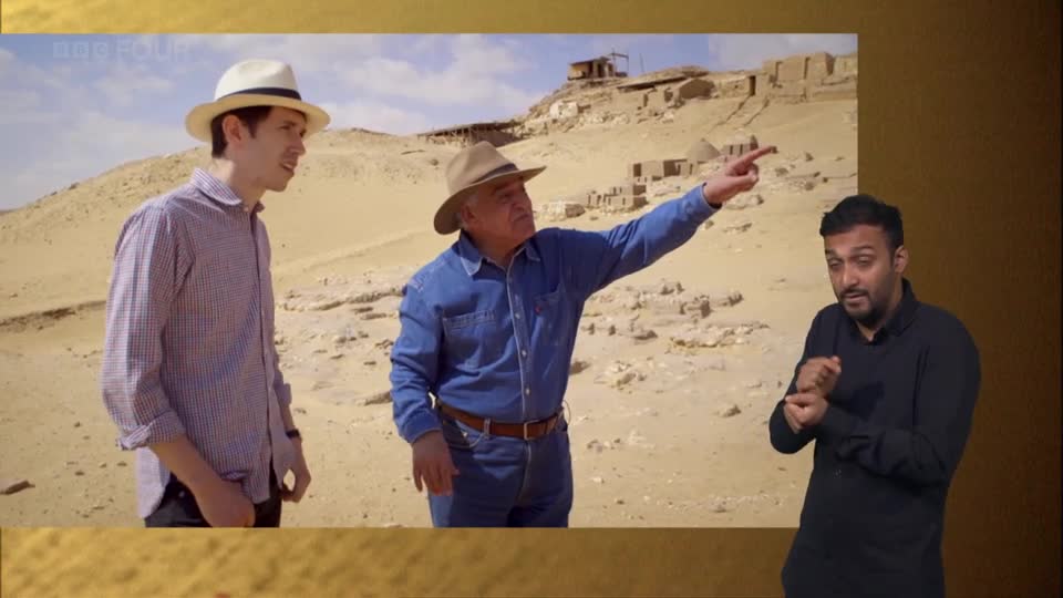 A man in a denim shirt points toward ancient structures in the distance, while another man in a checkered shirt looks on. A third man, wearing a black shirt, signs with his hands to the right of the screen.