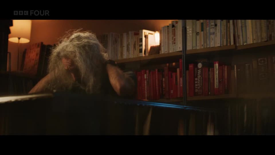 A person with long, unkempt grey hair leans over a desk, their face obscured. Behind them, shelves packed with books stretch across the wall, illuminated by a warm lamp.