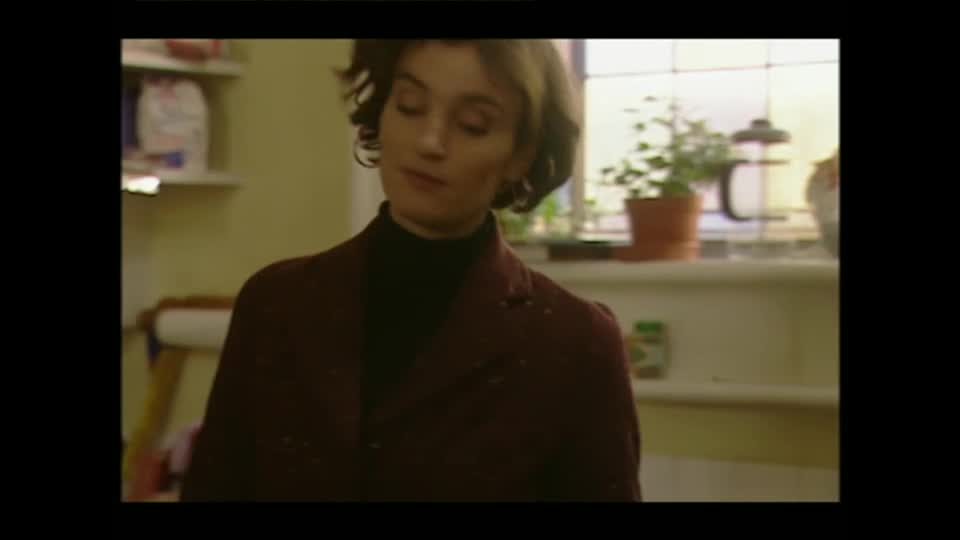 A woman in a dark coat stands in a kitchen, her head tilted slightly. Sunlight streams through a window behind her, illuminating a potted plant.