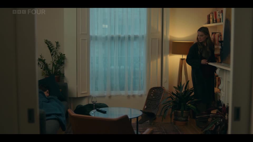 A woman in a dark robe stands by a fireplace, looking at a television. A person is tucked under a blue blanket on a sofa to the left.