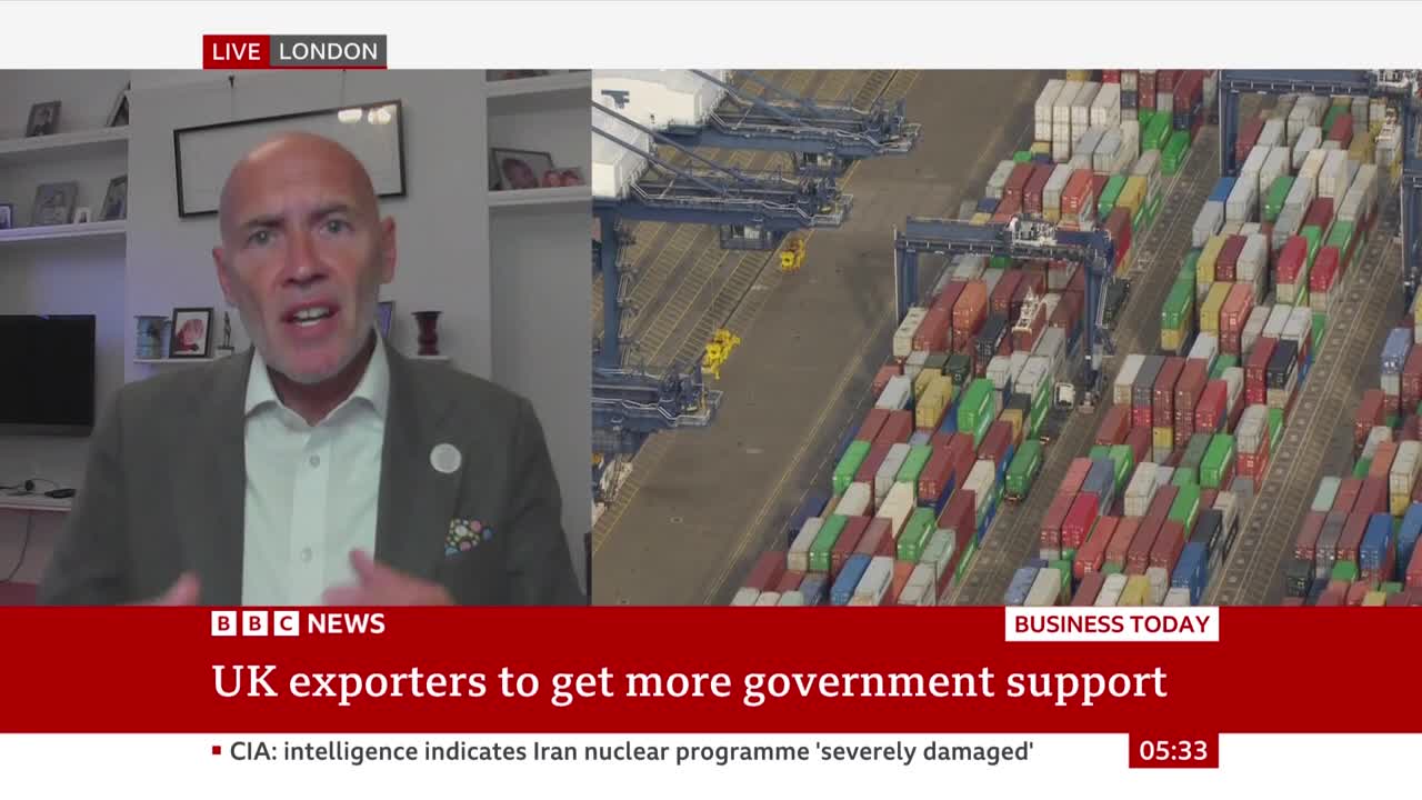 A man in a blazer gestures emphatically while speaking on BBC News from London. Beside him, a shot of a busy port shows rows of colorful shipping containers.
