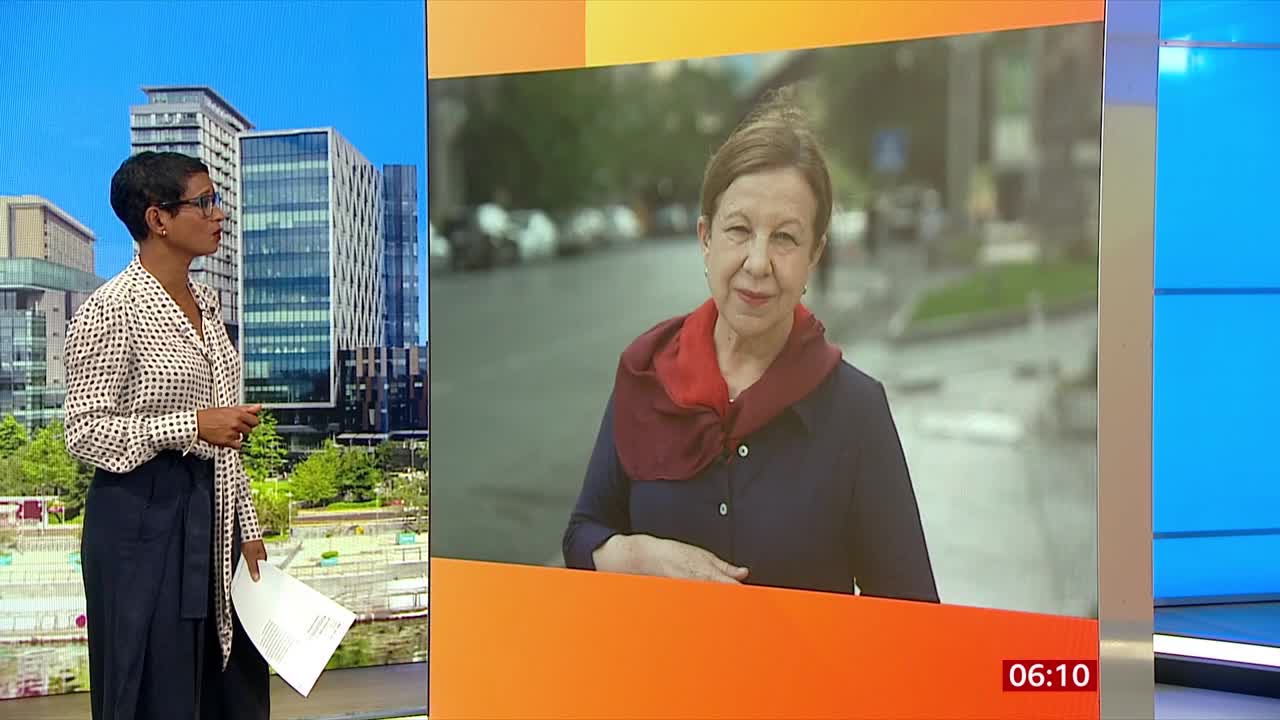 On the BBC News set, a presenter stands facing a screen. A woman in a red scarf and dark top appears on the screen, likely reporting from the United Kingdom.
