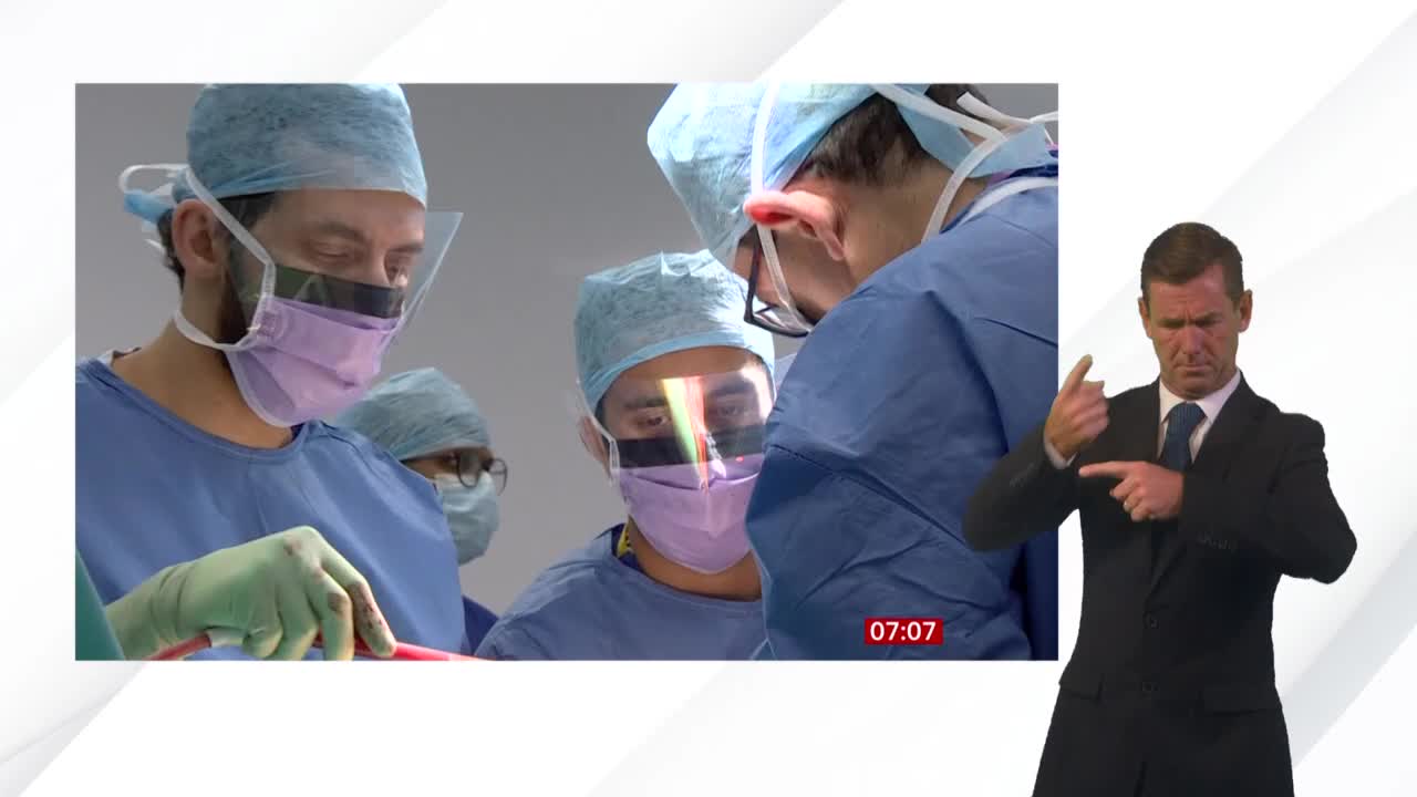 Inside a hospital operating room, several surgeons in blue scrubs and masks are focused on a procedure. A BBC News presenter in a suit is signing, presumably translating the medical update.
