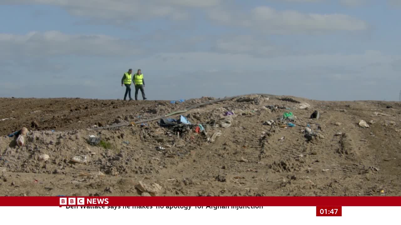 Two figures in reflective vests walk along the crest of a dirt mound, the landscape littered with debris. The BBC News logo sits at the bottom of the screen, with a headline about an apology.
