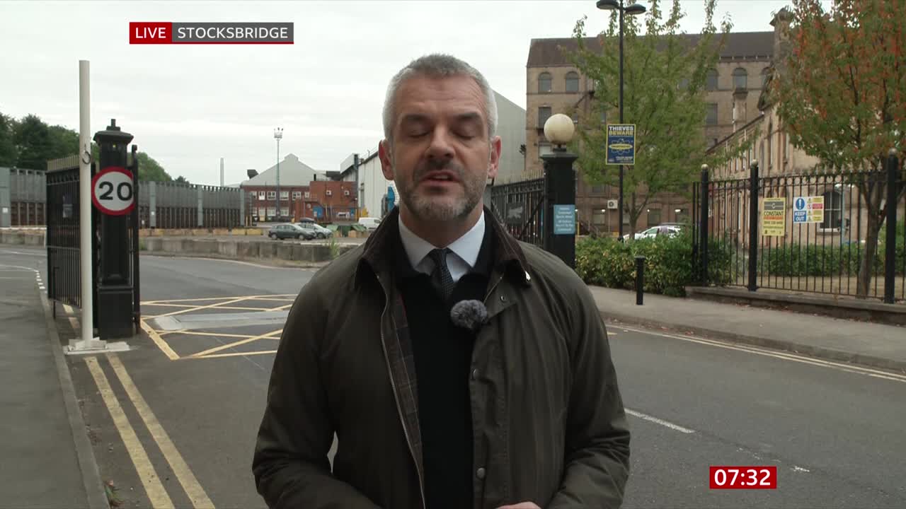 The BBC News reporter, standing on a paved road in Stocksbridge, is speaking directly to the camera. Behind him, a wrought-iron gate and a brick building are visible.
