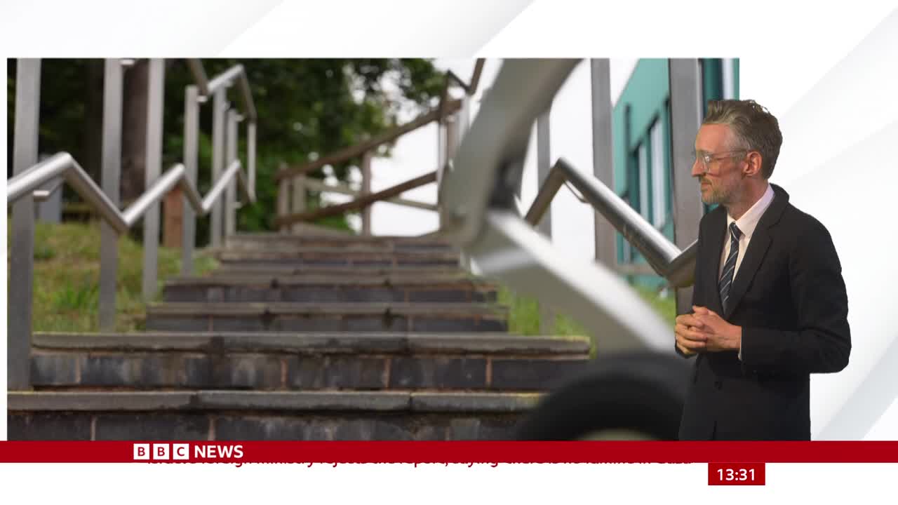 A news presenter in a suit stands to the side of a set of outdoor stairs. The camera focuses on the steps and the metal handrails, as if the location is part of the story.
